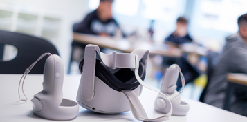 Im Vordergrund eine VR-Brille mit den Handsticks liegen auf dem Tisch, im Hintergrund sind Kinder erkennbar.